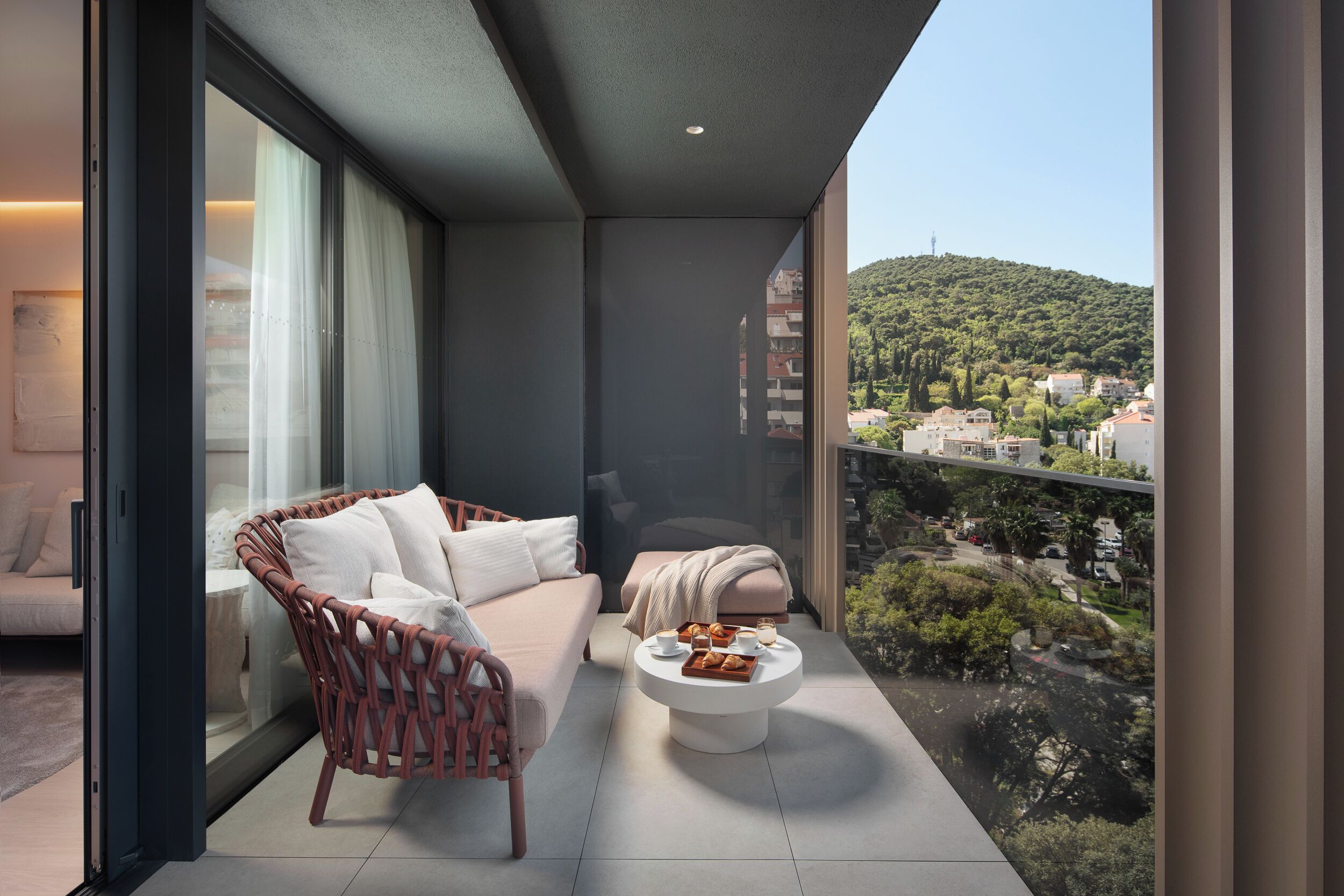 A balcony with a wicker chair, a white sofa, a round coffee table with pastries, and a city view.