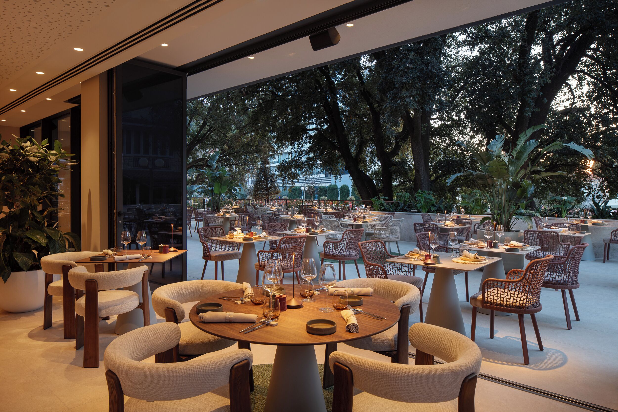Un área de comedor al aire libre con mesas y sillas redondas, rodeada de grandes ventanales y plantas en macetas.