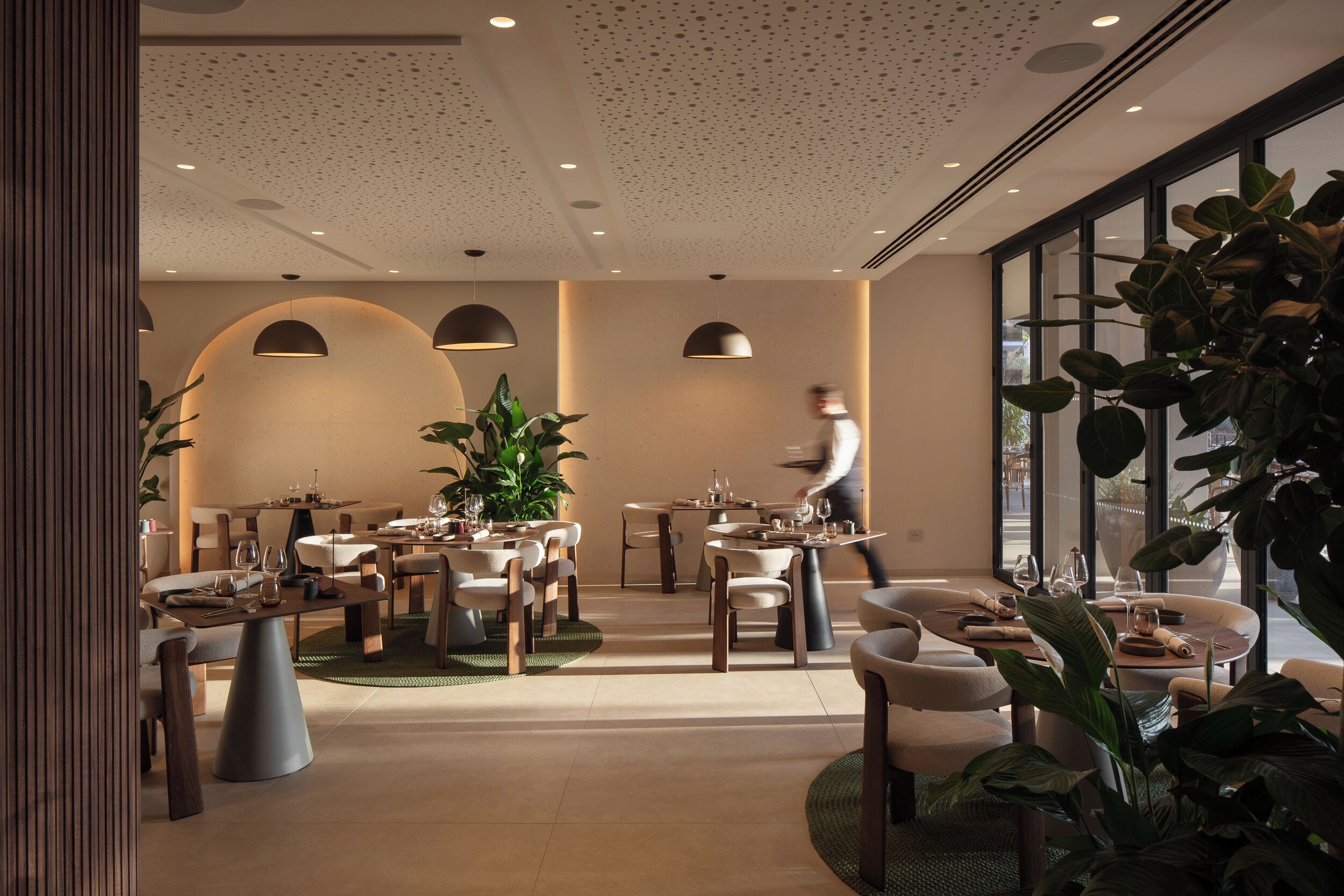 A modern dining area with multiple tables and chairs, large windows, and indoor plants.