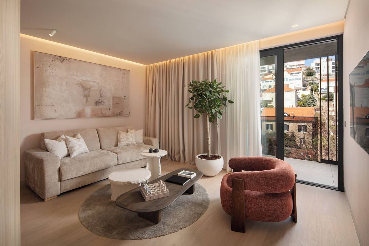 A modern living room with a sofa, coffee table, and a red chair. There is a large window with a view of buildings and a potted plant.