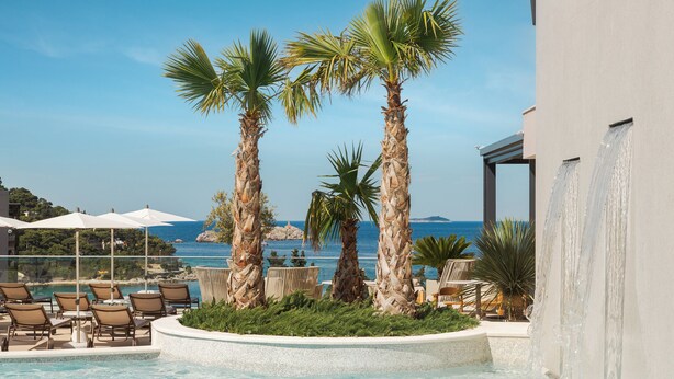 A pool area with lounge chairs, palm trees, and a view of the sea. - Hotel Sumratin (Dubrovnik)