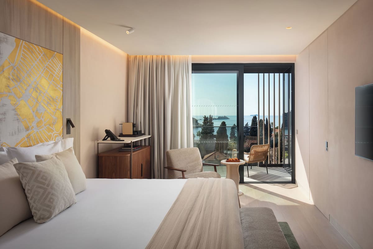 A modern hotel room with a large bed, a desk, a chair, a small table, and a balcony with a view.