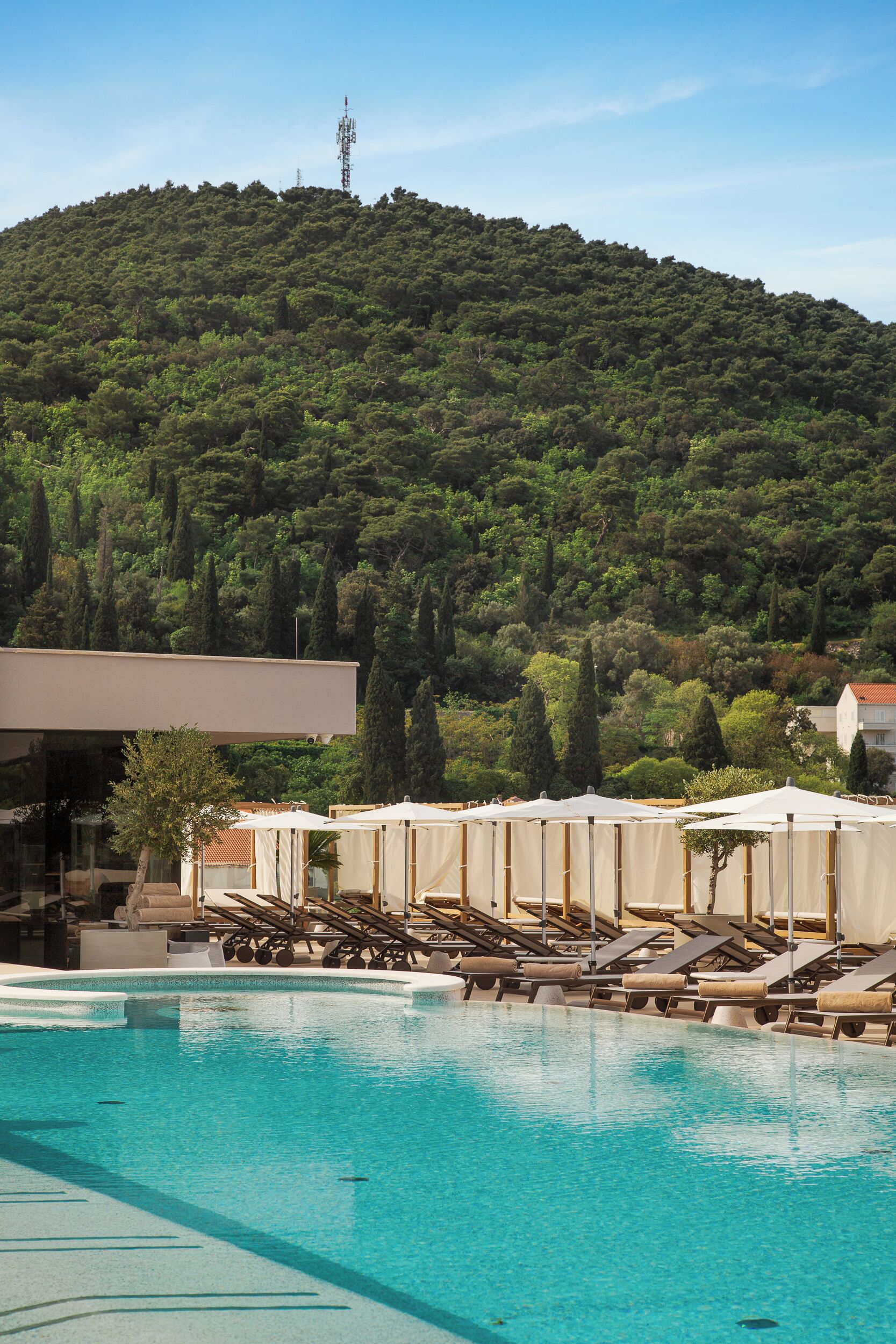 Una piscina con sillones de descanso y sombrillas, un edificio moderno, y una colina con árboles y una torre de comunicaciones.