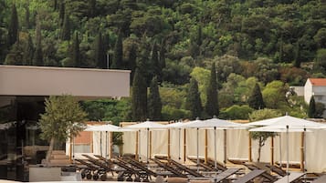 Una piscina con sillones de descanso y sombrillas, un edificio moderno, y una colina con árboles y una torre de comunicaciones.