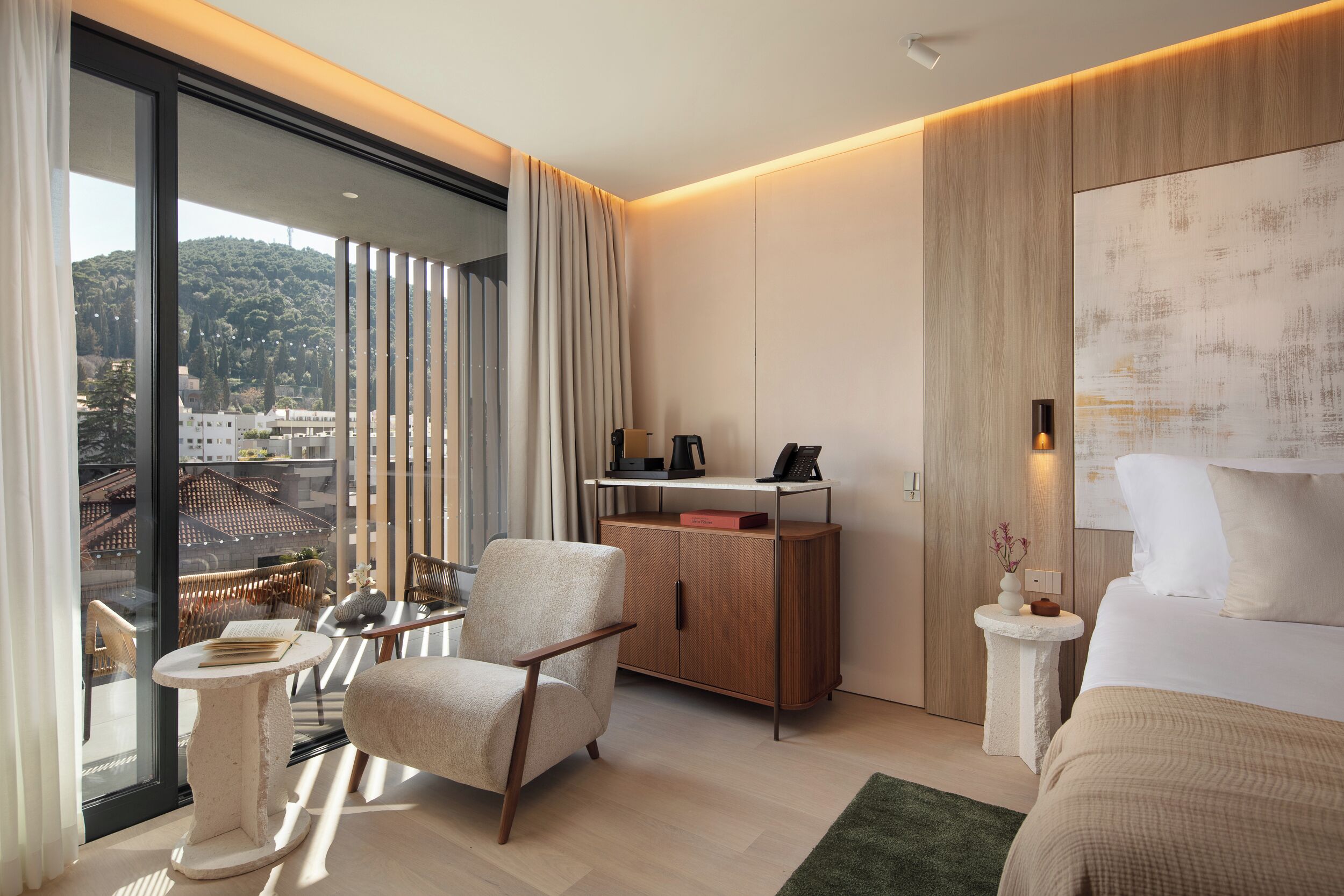 Quarto de hotel moderno com uma cama grande, área de estar com uma cadeira e mesa, um pequeno armário e vista para a cidade através de amplas janelas.