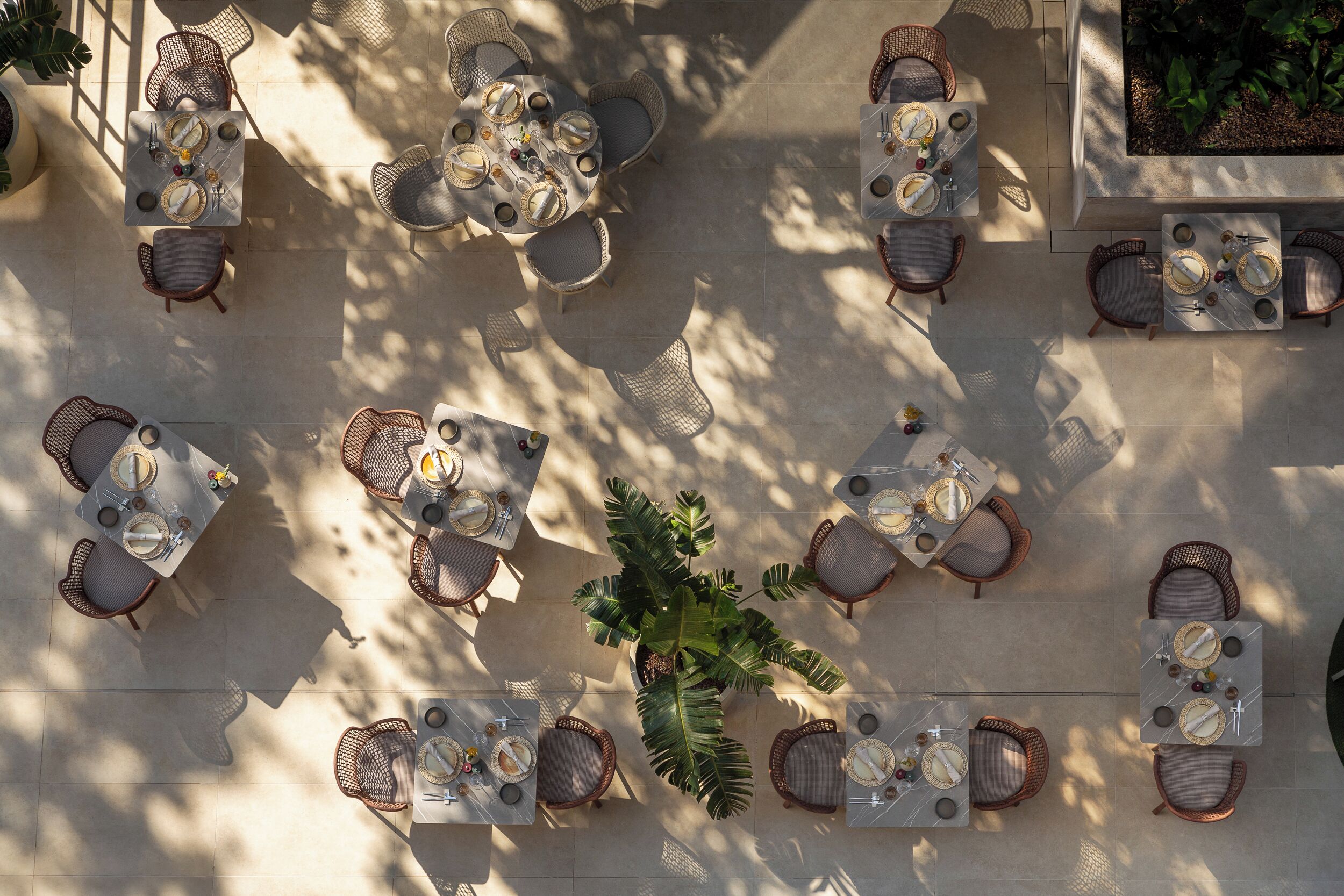 Een terras met ronde tafels en rieten stoelen, omgeven door potplanten.