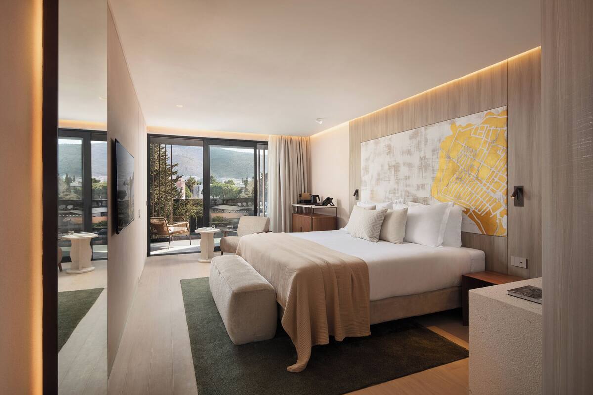 A modern hotel room with a large bed, a view of the outdoors, a map artwork on the wall, and a small side table.