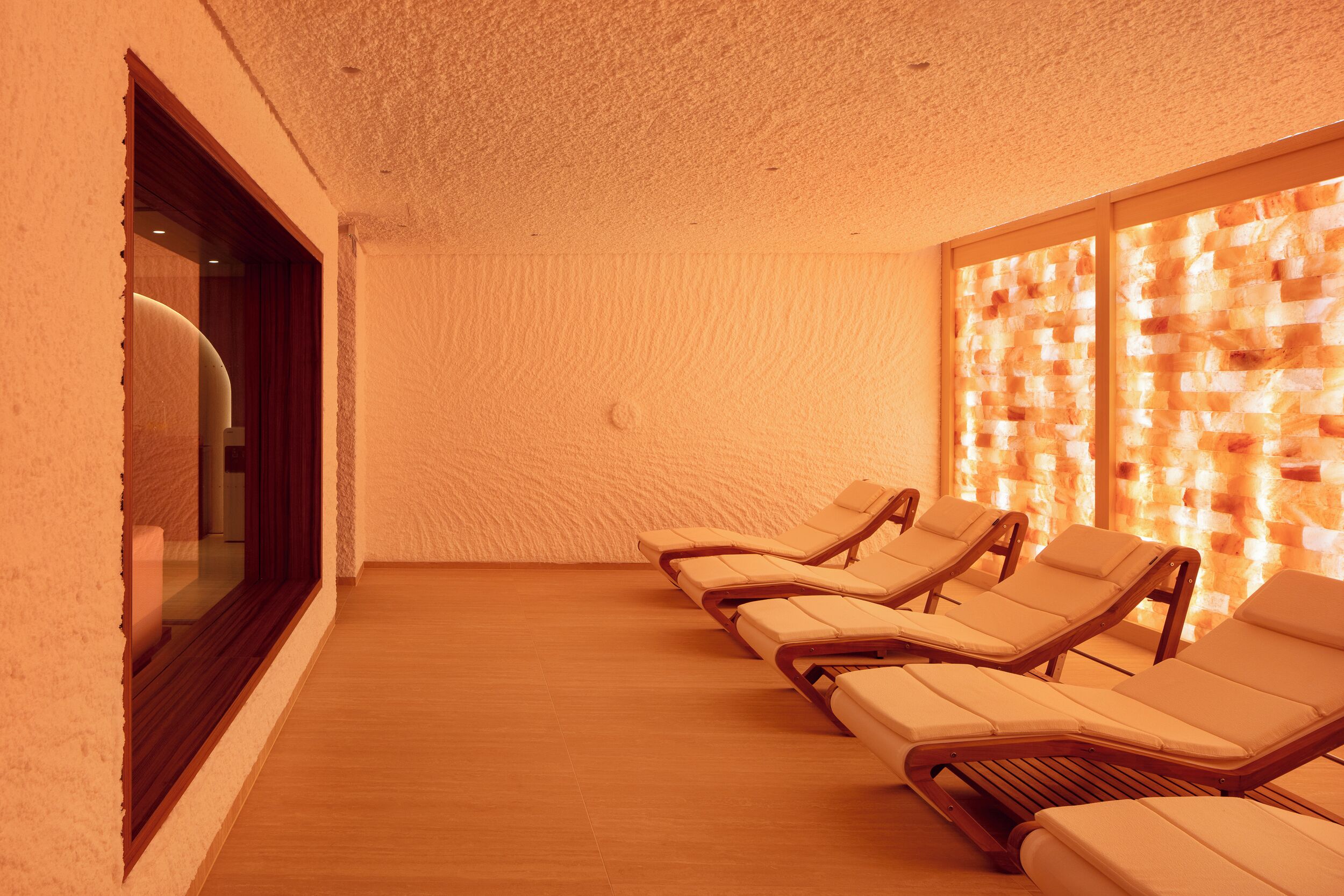 A spa room with wooden flooring, a row of sun loungers, a large window, and a textured wall.