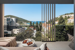 A balcony with a view of a coastal town, a dining table with pastries, and a bed with a view. - Hotel Sumratin (Dubrovnik)