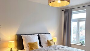 Une chambre avec un grand lit, une table de chevet avec une lampe, une fenêtre avec des rideaux et un luminaire suspendu.