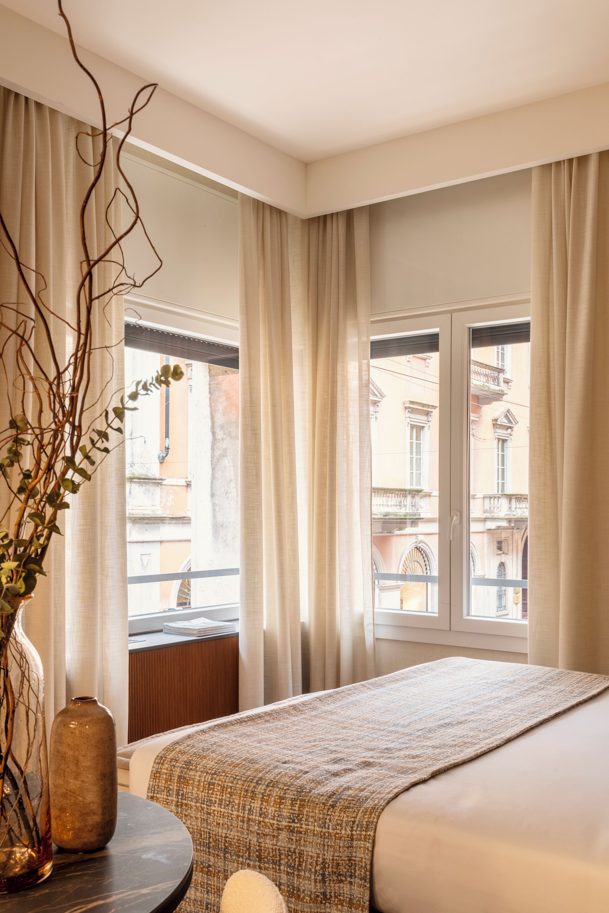 Habitación de hotel con cama, una mesa redonda con un florero y vista a edificios a través del ventanal.