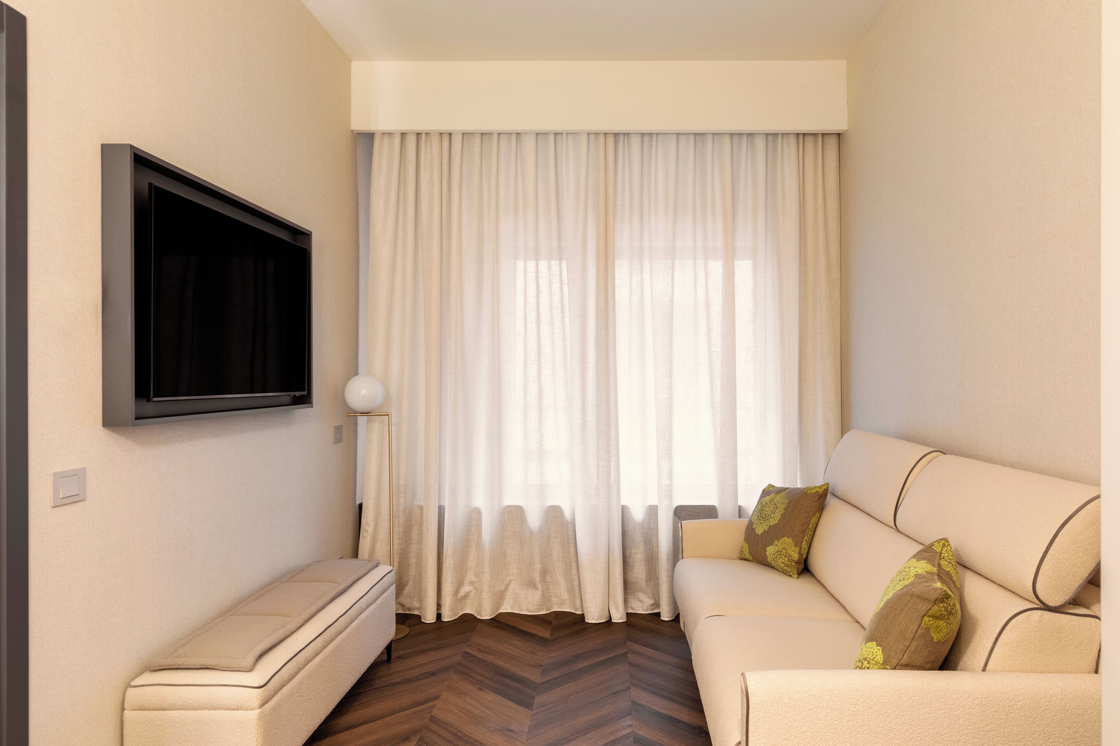 A living room with a sofa, a flat-screen TV mounted on the wall, and a large window with sheer curtains.