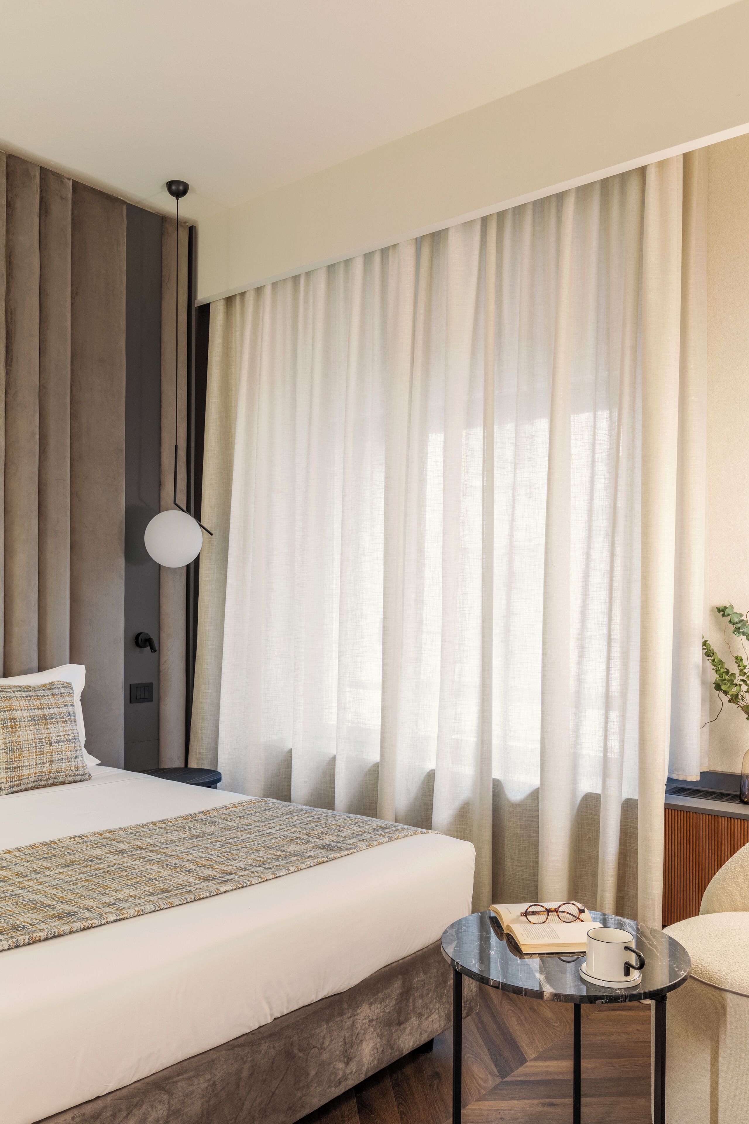 A hotel room with a large bed, a bedside table with a book and glasses, and a view of the outdoors through sheer curtains.