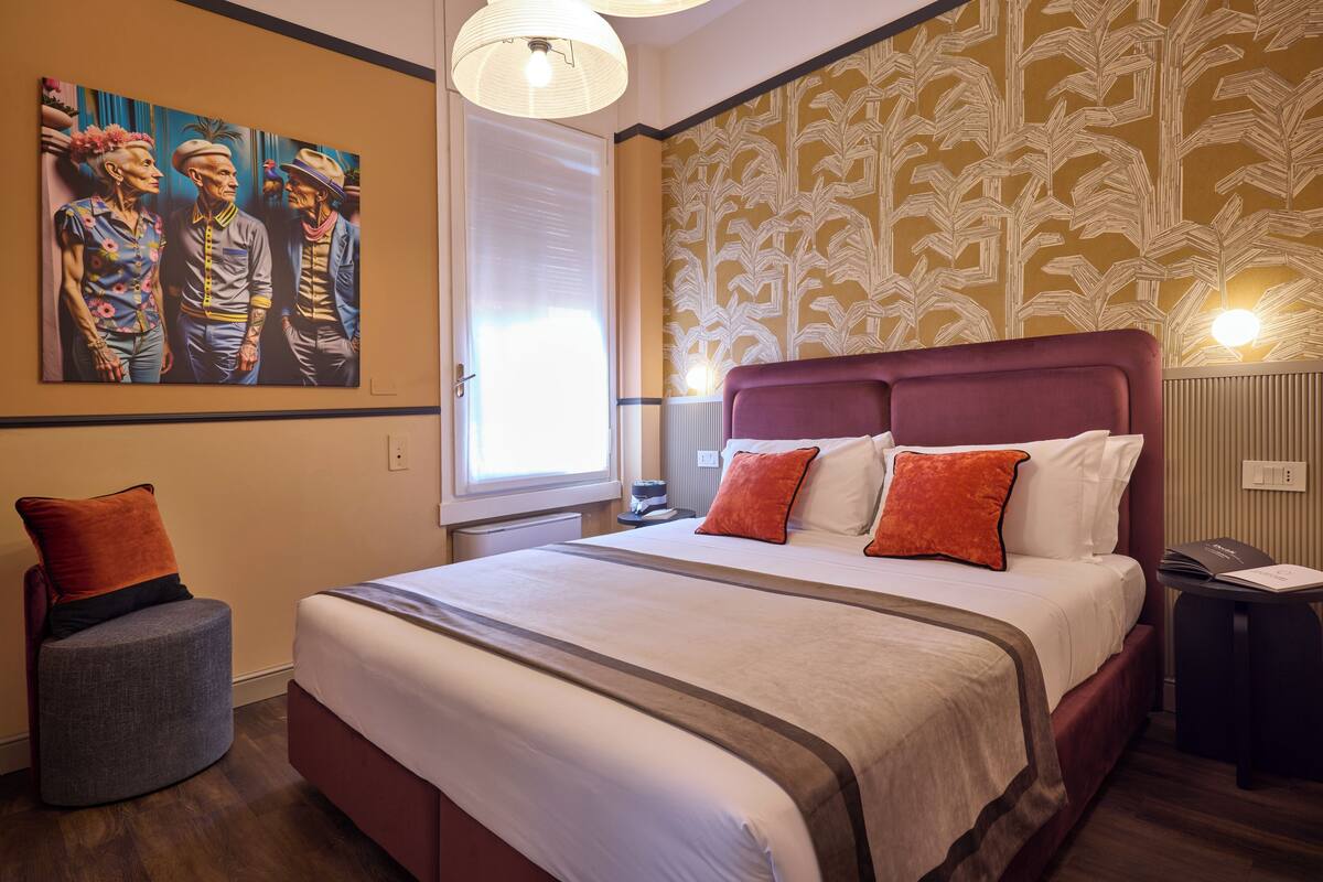 Un dormitorio con una cama grande, una mesita de noche, una silla, un cuadro en la pared y una ventana con persianas.