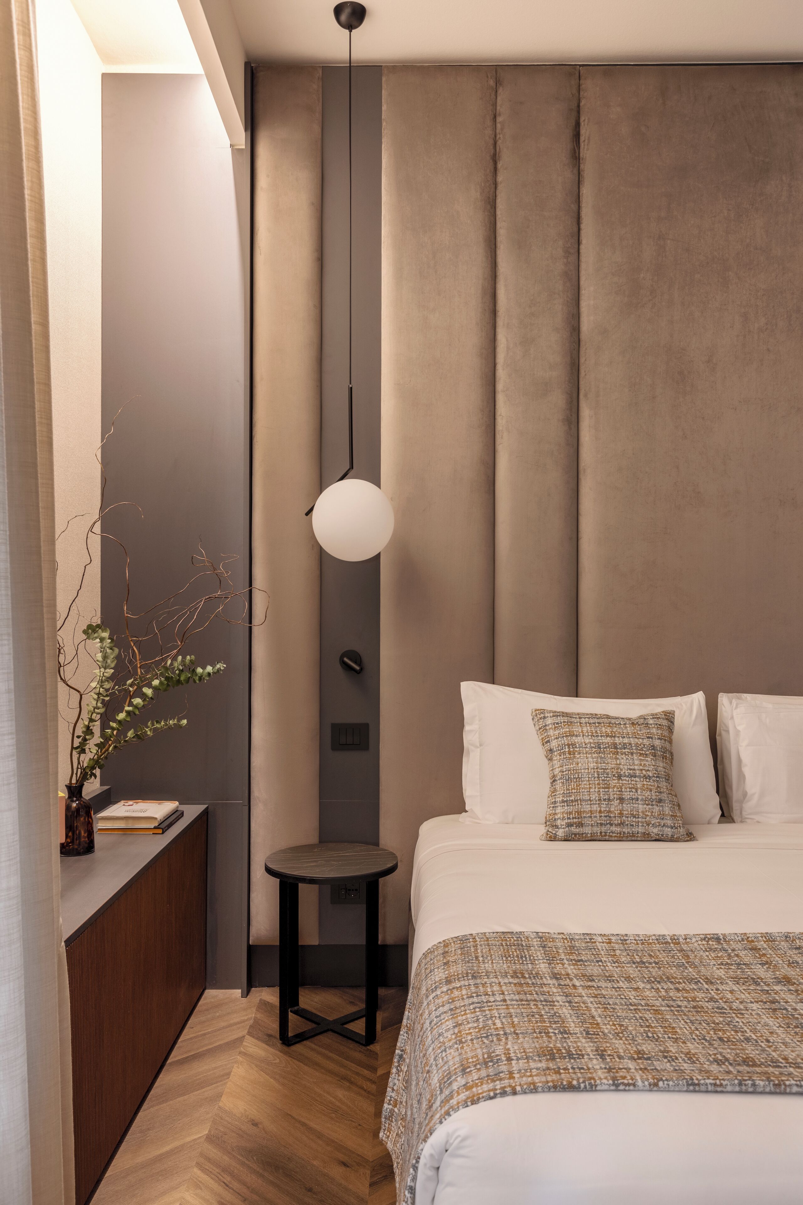 Een moderne slaapkamer met een groot bed, een nachtkastje, een klein krukje, een hanglamp en een plant.