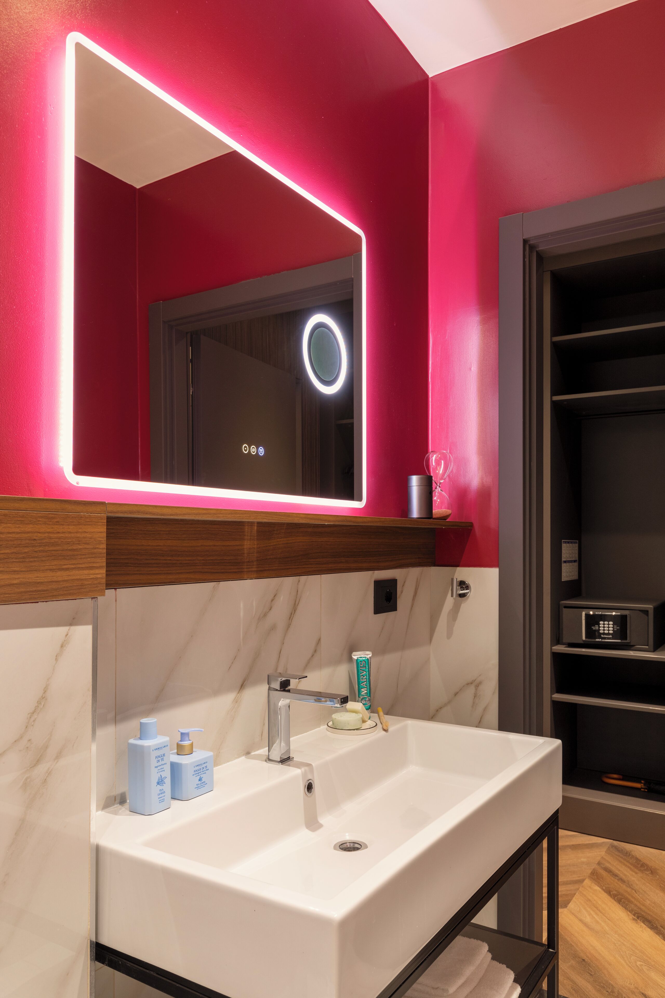 Ein Badezimmer mit einem weißen Waschbecken, einem Spiegel mit Neonbeleuchtung und einem Holzregal.