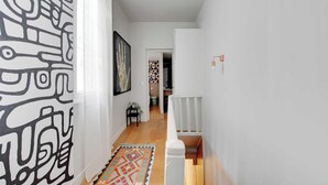 Un couloir avec un mur orné de motifs, un tapis, une œuvre d’art encadrée et une rambarde blanche.