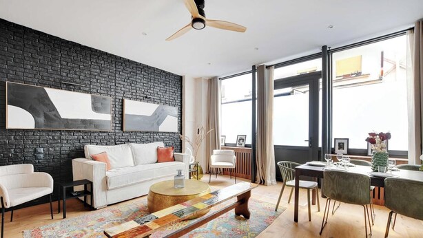 Un salon moderne avec un mur en briques noires, un canapé blanc, une table basse ronde et un coin repas avec une table et des chaises vertes.