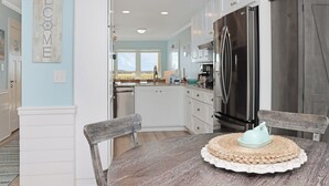 A modern kitchen with white cabinetry, a stainless steel refrigerator, and a wooden dining table with a placemat. - 🌊Dog Friendly-Steps2Bch-Beach Views-Sea Cliff A5 Sunset Shores (Cape San Blas)