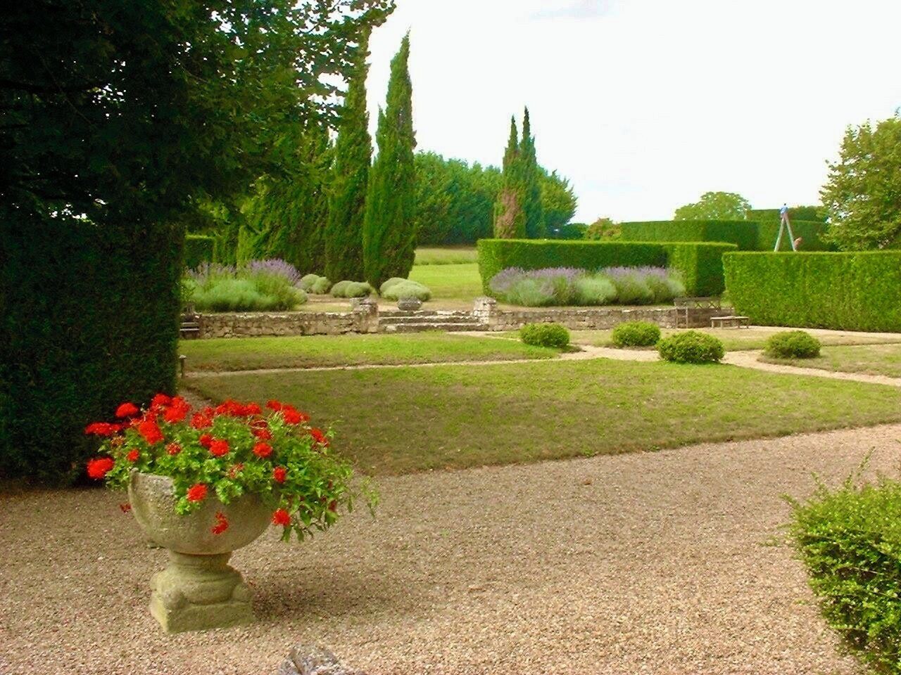 Un jardín formal con setos cuidadosamente recortados, un camino de grava y una jardinera de piedra con flores rojas.