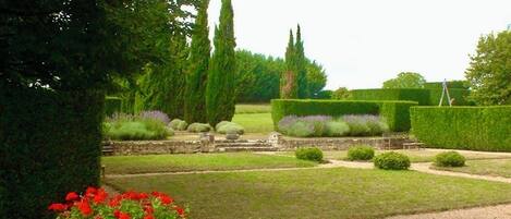 Un jardín formal con setos cuidadosamente recortados, un camino de grava y una jardinera de piedra con flores rojas.