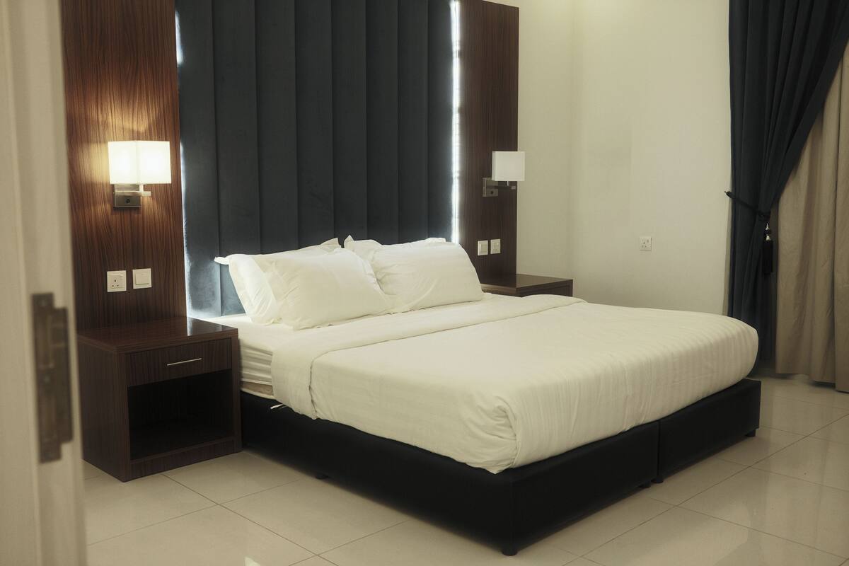 Une chambre d’hôtel moderne avec un grand lit, une tête de lit en bois foncé, des tables de chevet avec des lampes et une grande fenêtre avec des rideaux.