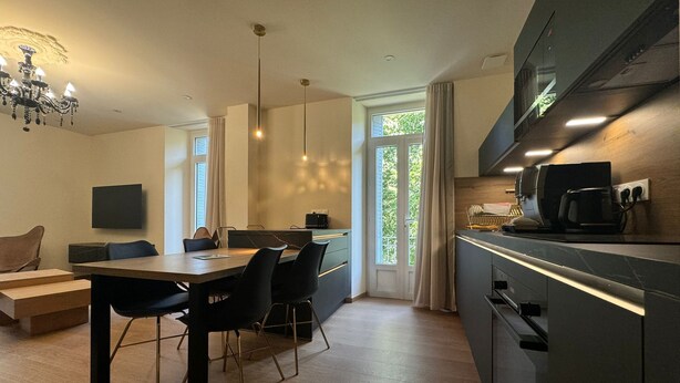 A modern kitchen with a dining area, featuring a chandelier, a flat-screen TV, and a large window with curtains. - Residence VillaBelleFleur-Apartment La Colchique (Château-Ville-Vieille)
