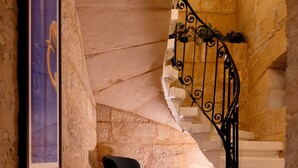 Una escalera de caracol con barandilla de metal negro, un cuadro enmarcado en la pared y una silla negra en un pasillo de paredes de piedra.