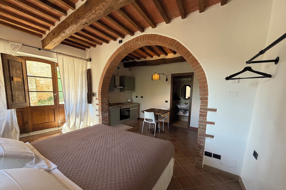 Een slaapkamer met een bed, een houten deur, een keuken met een eettafel en stoelen, en een boogvormige bakstenen constructie.