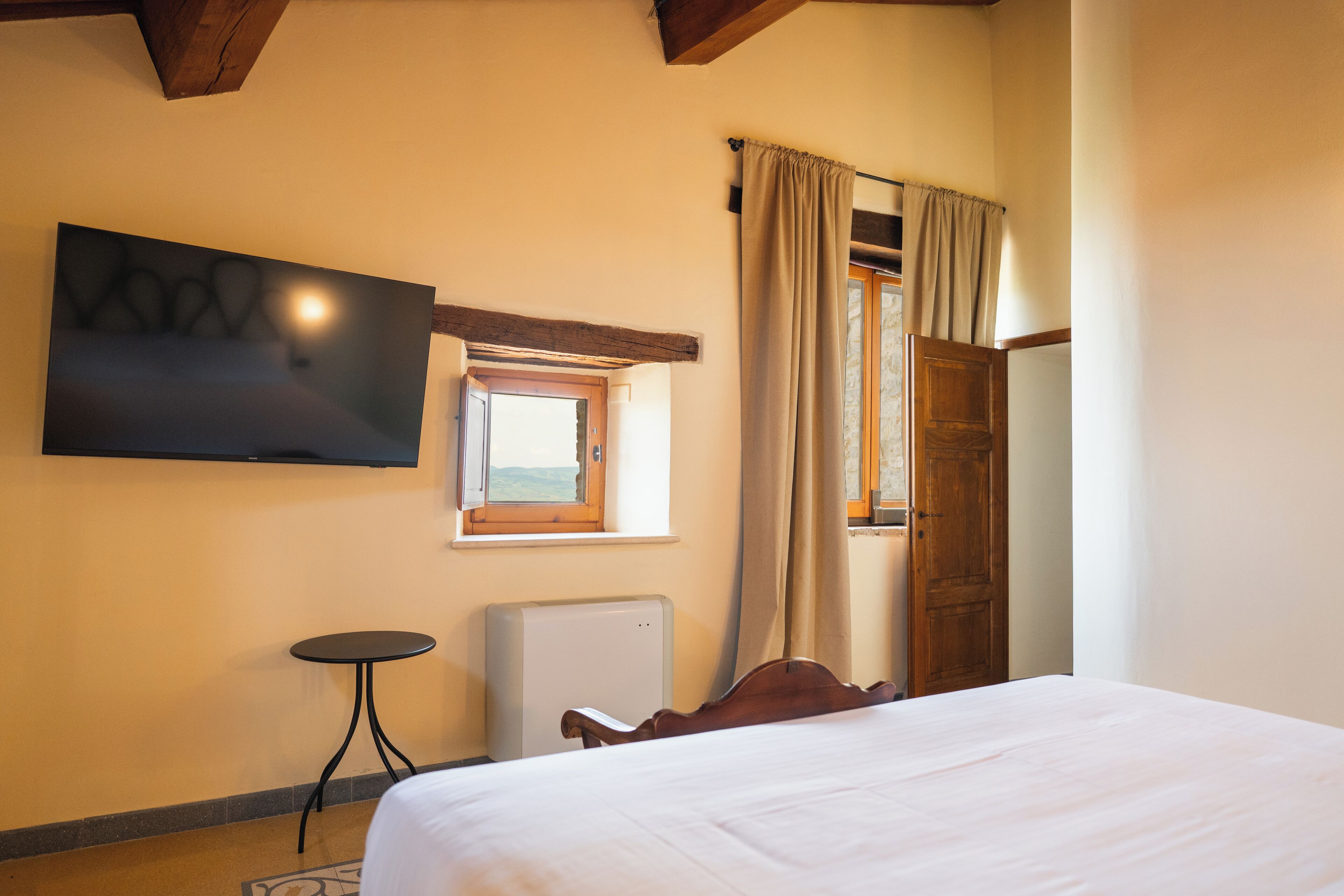 A hotel room with a bed, a TV mounted on the wall, a small table, a window with curtains, and a door.
