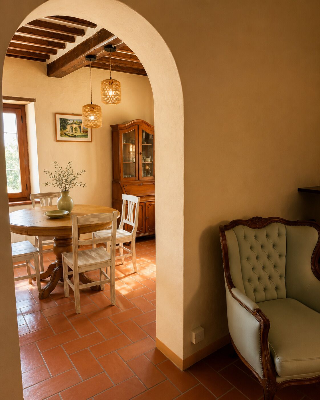 A dining area with a round table, chairs, a vase with flowers, a wooden cabinet, and a cushioned armchair.