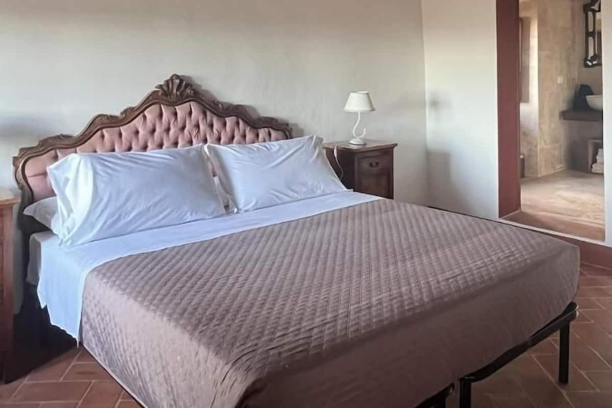 Een slaapkamer met een groot bed, houten balken in het plafond, een nachtkastje met een lamp, en een badkamer die door een open deur zichtbaar is.