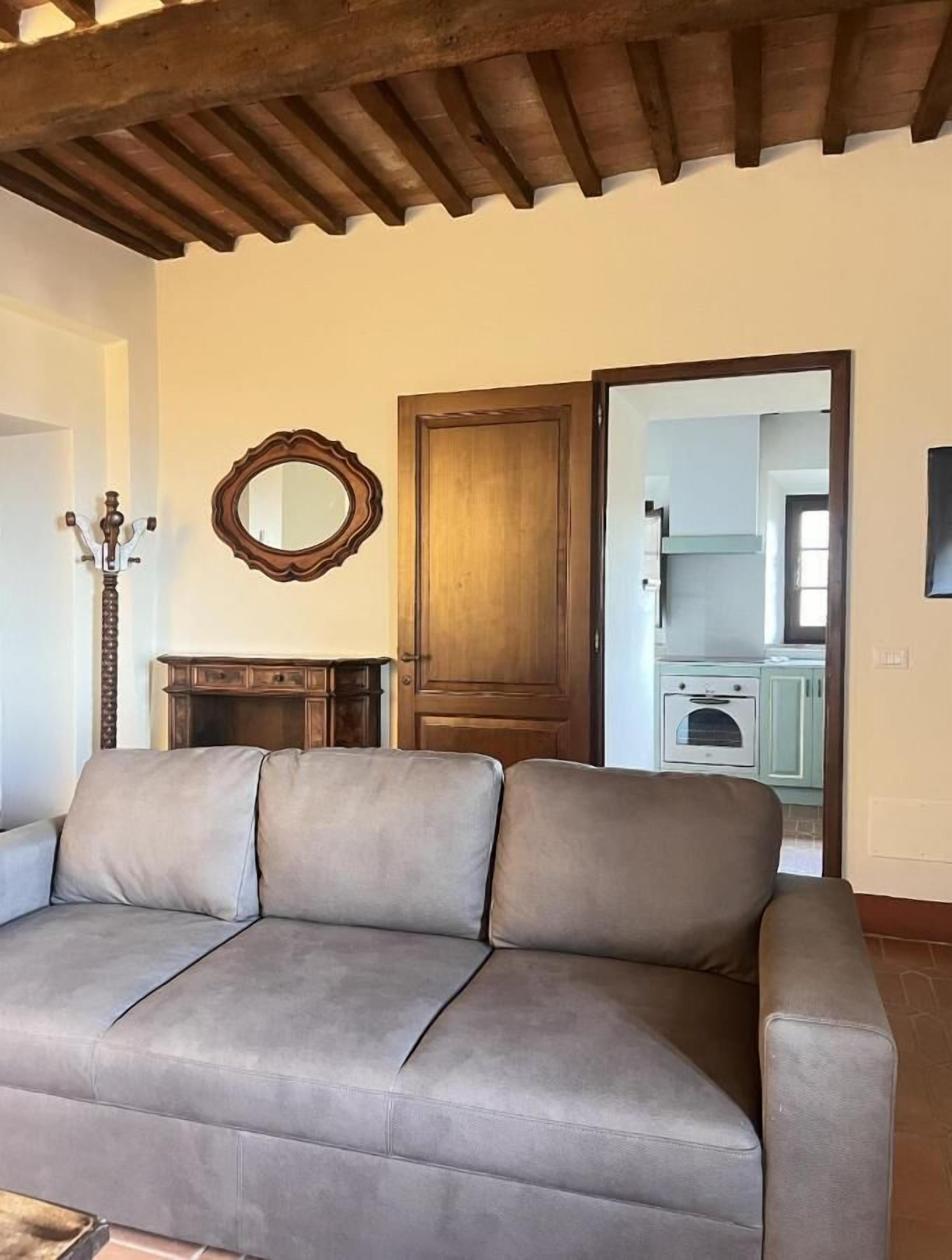 Una sala de estar con un sofá gris, vigas de techo de madera, una puerta de madera, un espejo y un aparador.