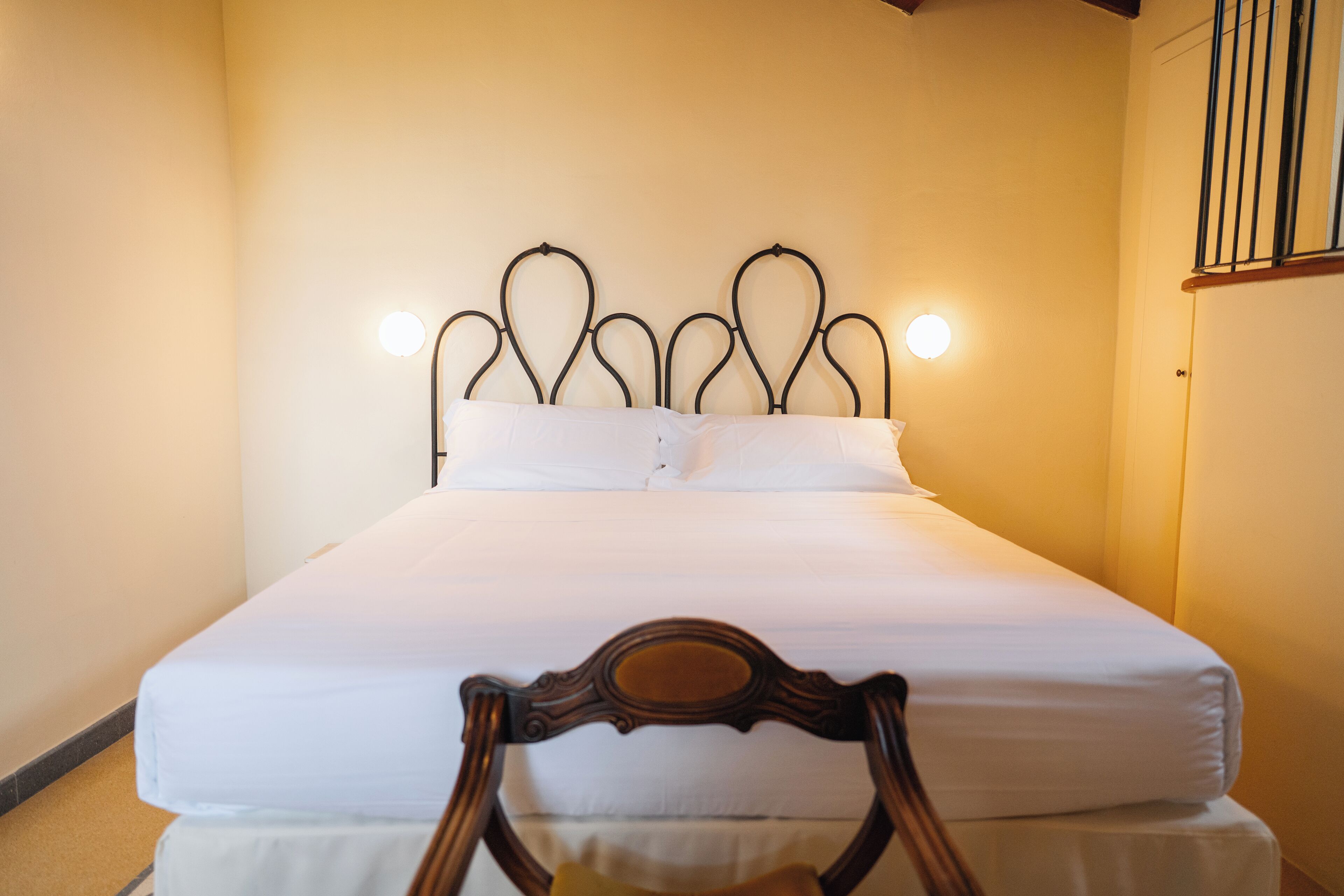 Una cama bien hecha con cabecera de metal, una silla de madera al frente y una lámpara colgante arriba.