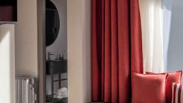A hotel room with a bed, a window with red curtains, a view of a bathroom, and a suitcase on the floor.