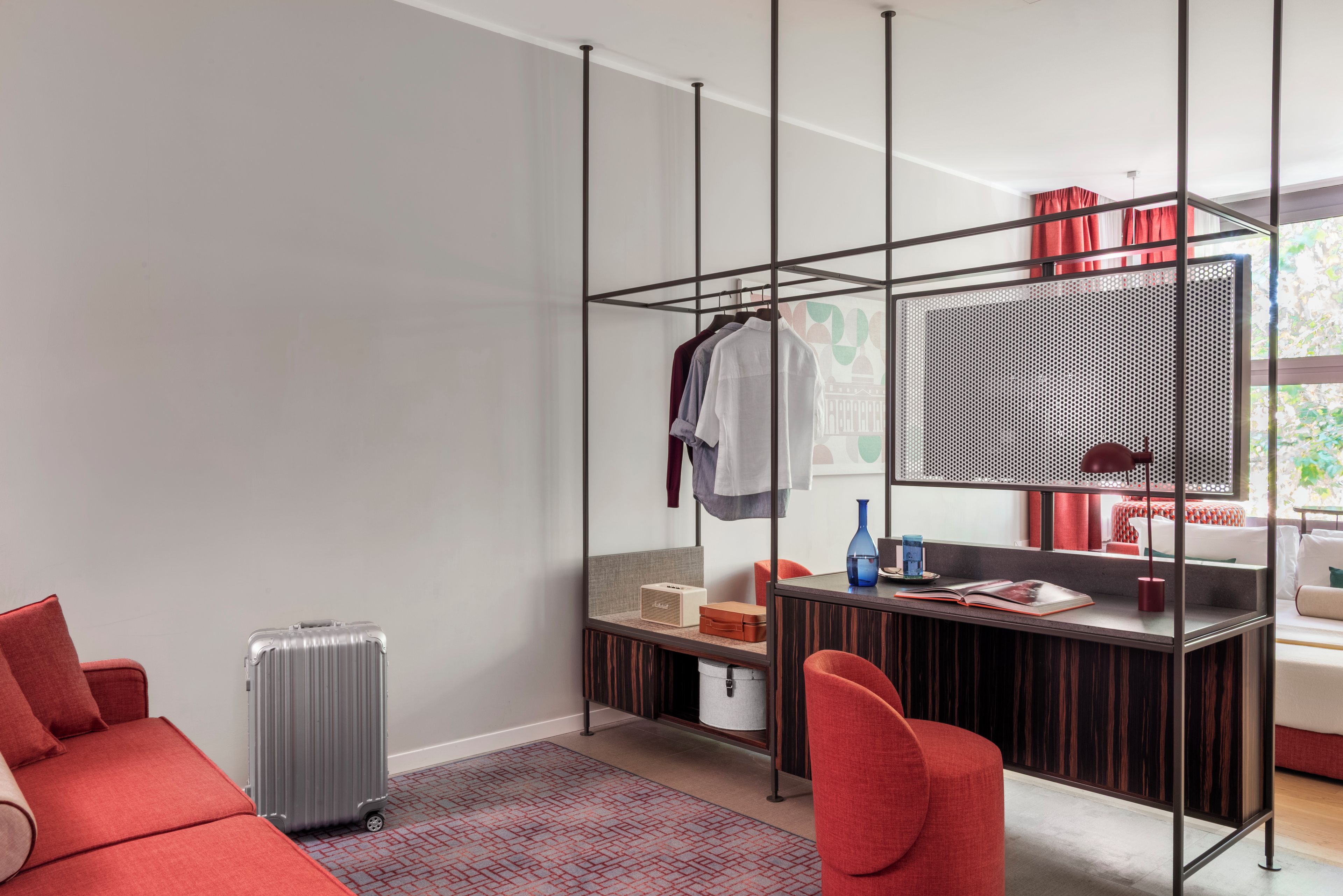 Ein modernes Zimmer mit einem roten Sofa, einem kleinen Schreibtisch mit Computer, einer Hängelampe und einem Koffer.