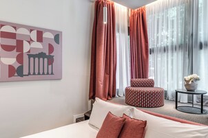 Quarto de hotel com uma cama, um banquinho estampado, uma pequena mesa com um vaso e um quadro na parede.