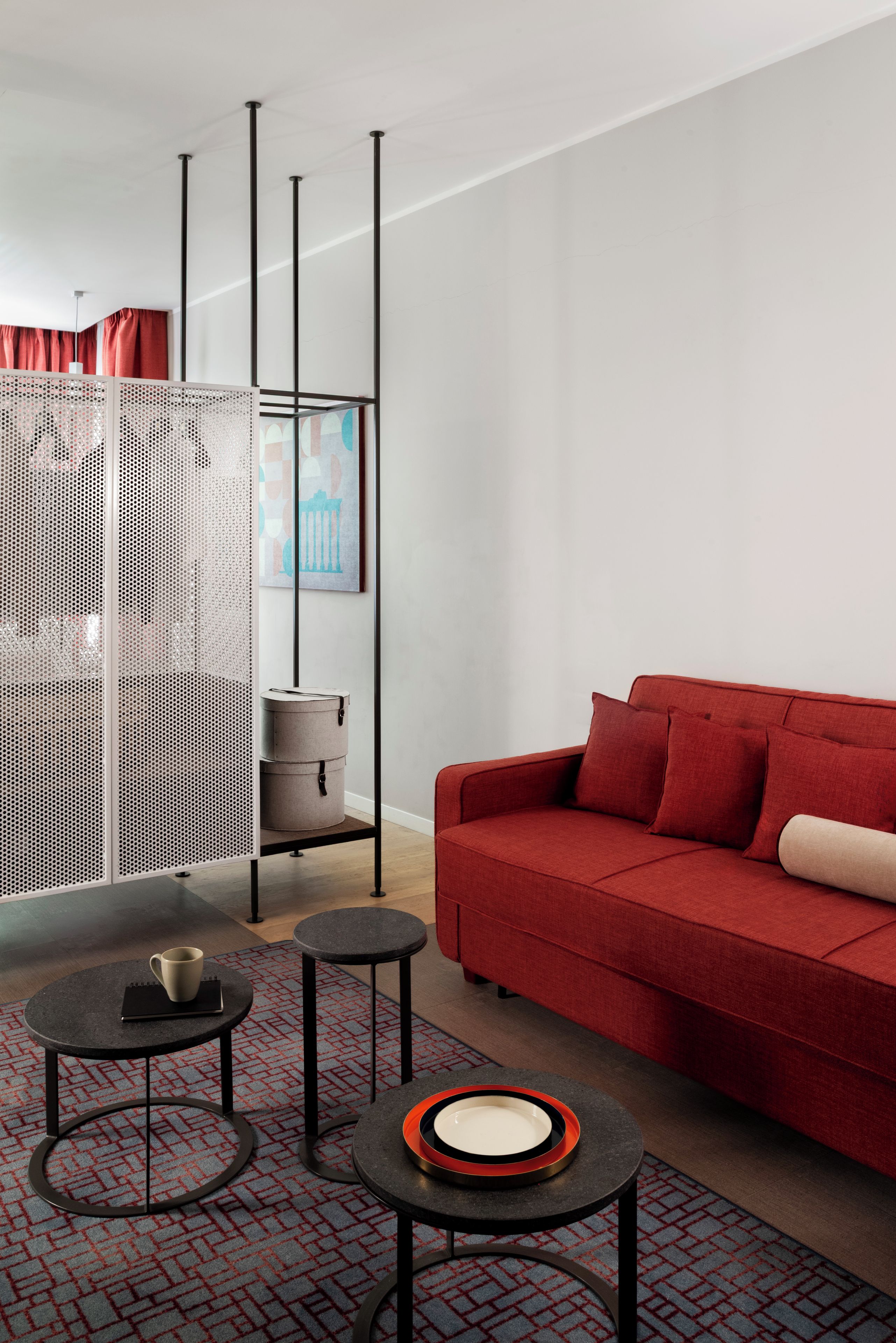 Un salon moderne avec un canapé rouge, deux petites tables rondes, un tapis à motifs et un panneau décoratif mural.