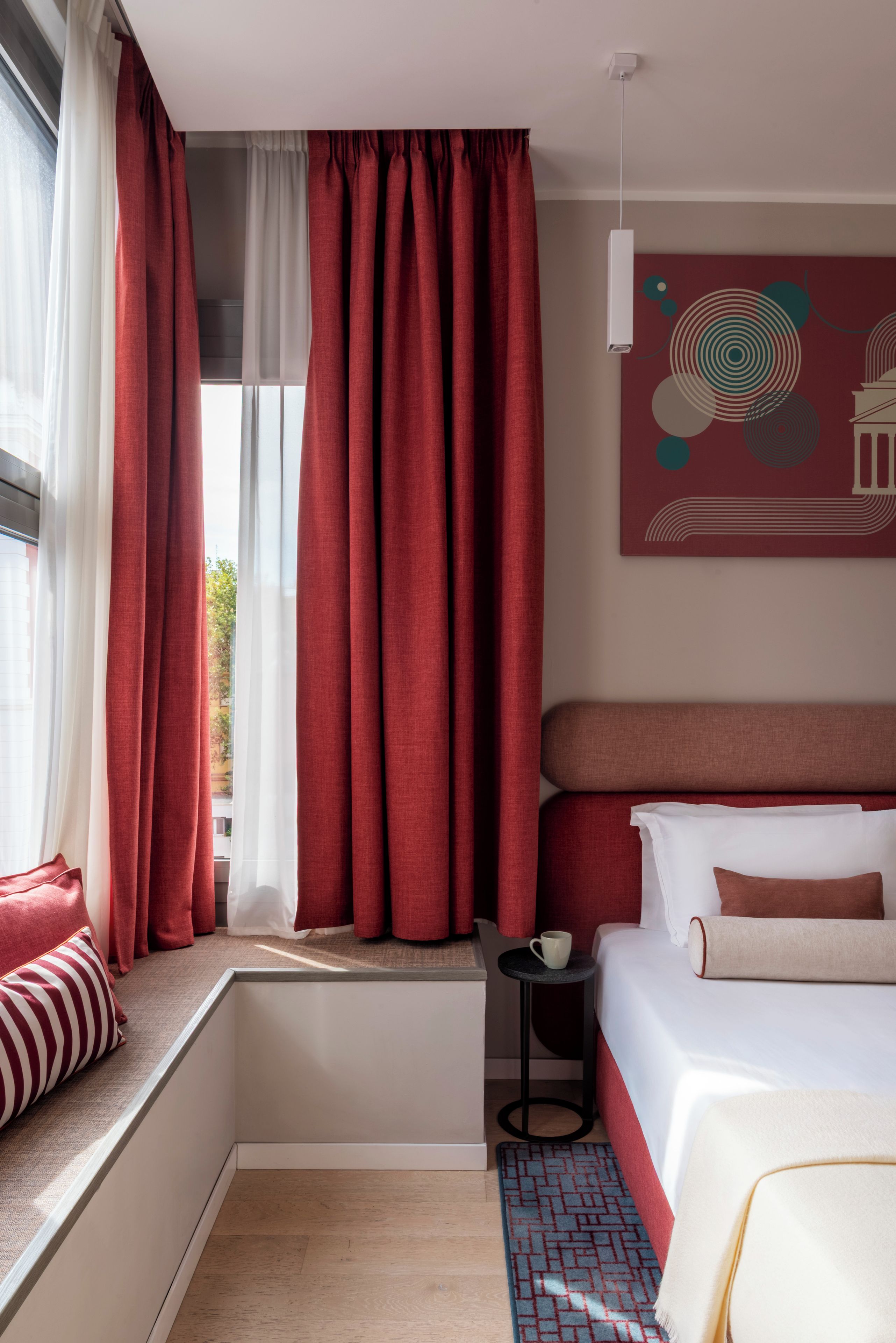 Habitación de hotel con cama, ventana con cortinas rojas, una mesita con una taza y una obra de arte en la pared.