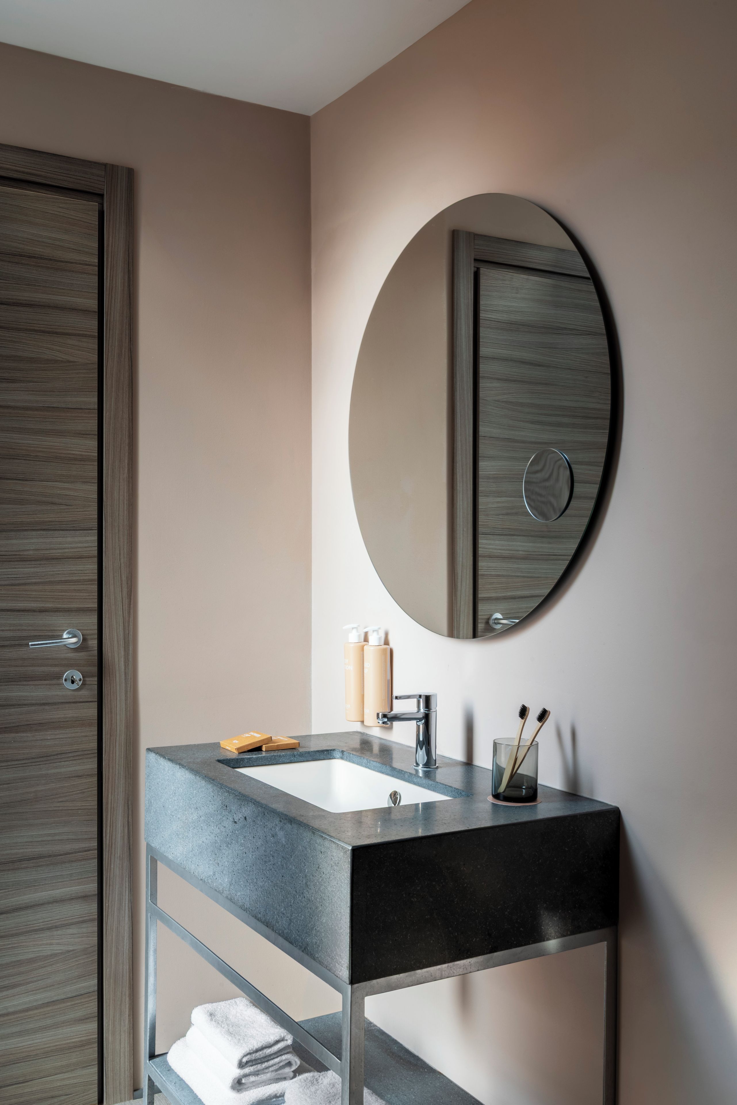 A modern bathroom with a wooden door, a round mirror, a sink with a faucet, and a towel rack.