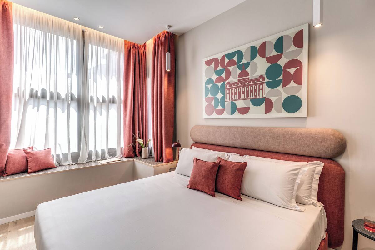 A modern hotel room with a large bed, a window with curtains, a wall-mounted artwork, and a bedside table with a lamp.