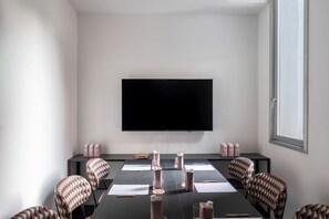 Een vergaderzaal met een groot flatscreen aan de muur, een lange tafel met papieren, pennen en fruitsapverpakkingen, en gestreepte stoelen.