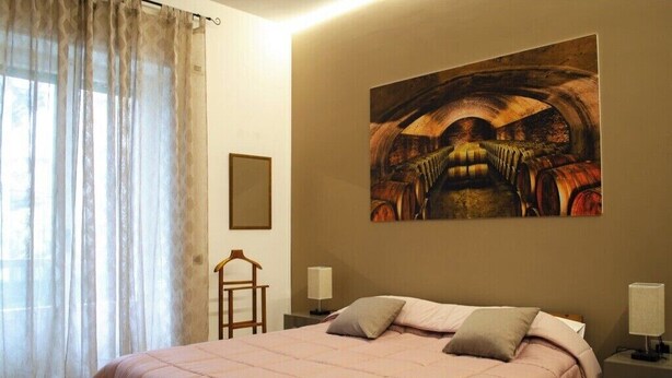 Ein Schlafzimmer mit einem großen Bett, einem Gemälde an der Wand, einem Nachttisch und einem Fenster mit transparenten Vorhängen.