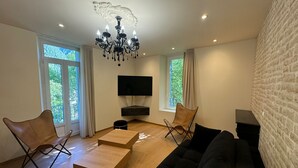 A modern living room with a black sofa, two leather chairs, a wooden coffee table, a TV mounted on the wall, and a chandelier. - Residence VillaBelleFleur-Apartment La Centaurée (Château-Ville-Vieille)