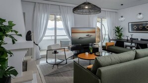 Ein modernes Wohnzimmer mit großem Flachbildfernseher, bequemen Sitzgelegenheiten, einem Essbereich und großen Fenstern mit Vorhängen.