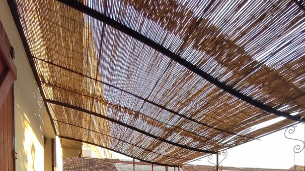 Un patio cubierto con vistas al campo, que incluye una mesa con un frutero, una botella y vasos, rodeado de sillones y plantas en macetas.