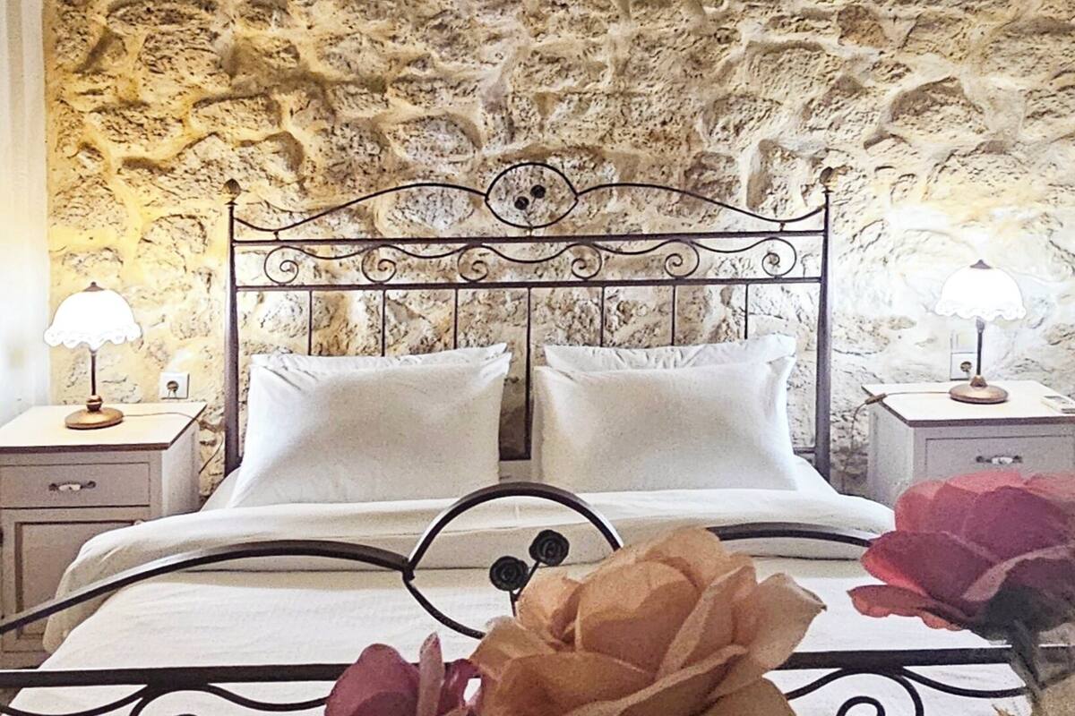 Un dormitorio con pared de piedra, estructura de cama de metal, ropa de cama blanca y una lámpara colgante.