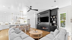 A modern living room with a fireplace, a large TV, and a dining area with white cabinetry and pendant lighting. - 🌴Almost Heaven • Gulf View Luxury with Private Heated Pool & Deeded Beach Access (Santa Rosa Beach)