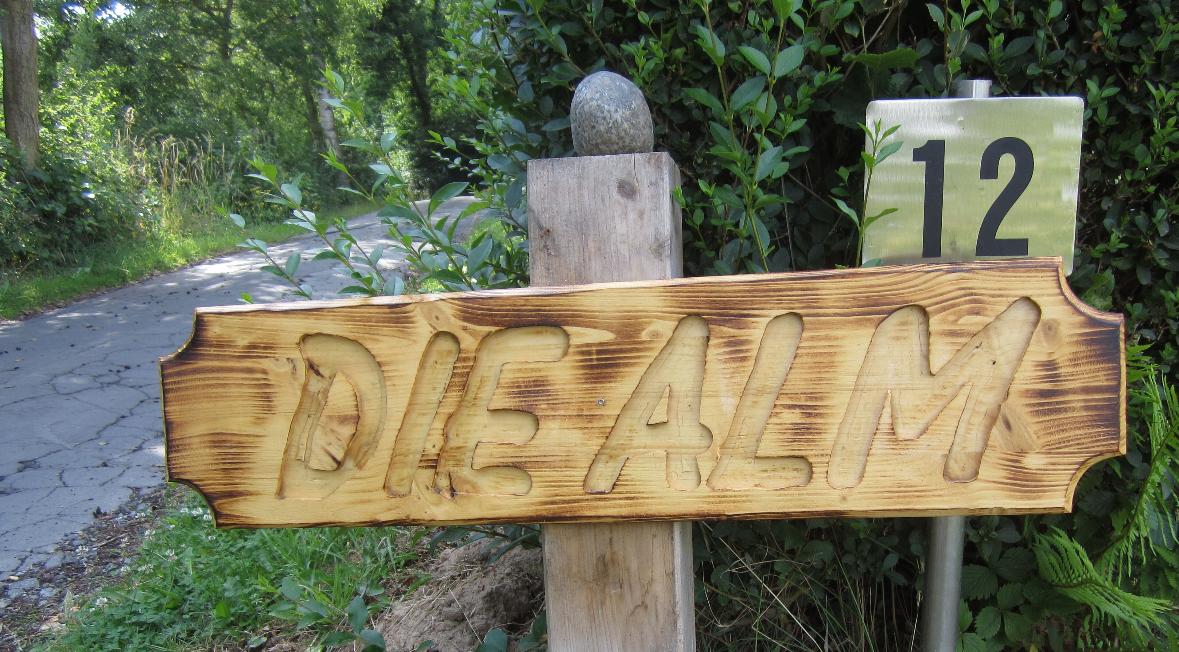 A wooden signpost with the name 'DIE ALM' carved into it, mounted on a post with a stone at the top.