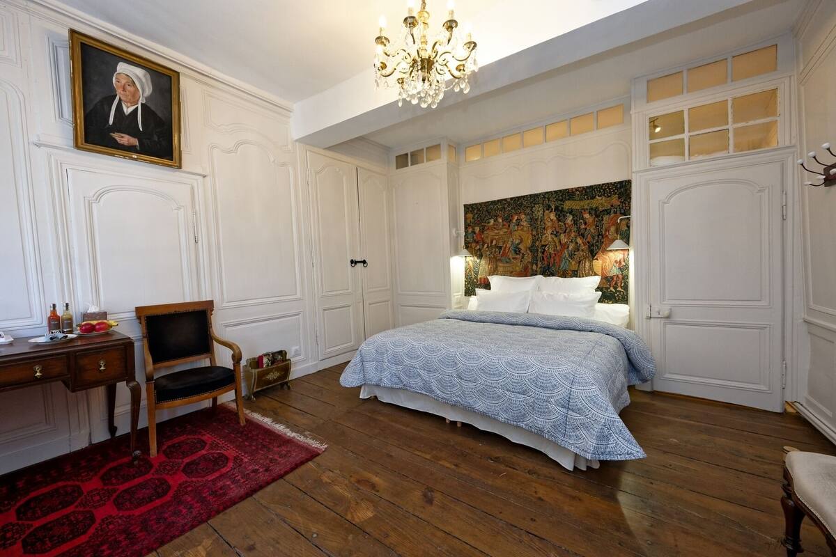 A bedroom with a large bed, a desk with a chair, a red rug, and a chandelier.